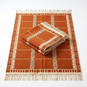 May include: A rectangular woven rug in burnt orange with cream-coloured geometric patterns and fringe on the short ends. A folded version of the same rug is placed on top, displaying the same pattern of vertical stripes and triangular motifs.