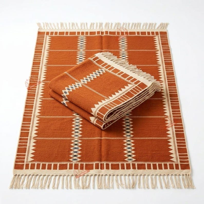 May include: A rectangular woven rug in burnt orange with cream-coloured geometric patterns and fringe on the short ends. A folded version of the same rug is placed on top, displaying the same pattern of vertical stripes and triangular motifs.