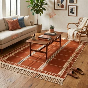 May include: A rectangular area rug featuring a burnt orange base with white geometric patterns, including zig-zag lines and grid-like borders, finished with long fringe on both ends. The rug is placed on a wooden floor beneath a wooden coffee table.