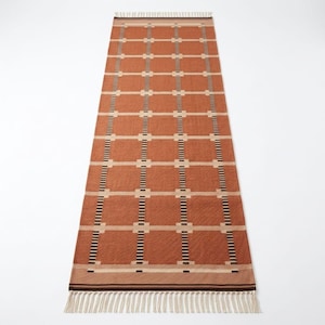 May include: A long, rectangular runner rug featuring a terracotta orange base color with a repeating grid pattern of cream-colored horizontal lines and vertical stripes composed of small, alternating black and white rectangular segments.