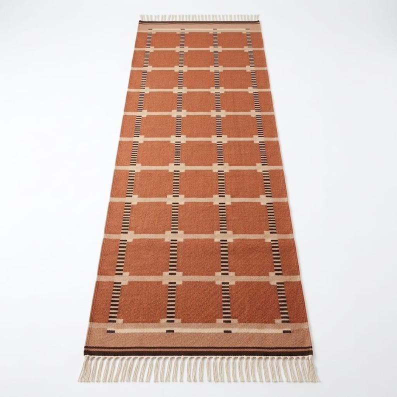 May include: A long, rectangular runner rug featuring a terracotta orange base color with a repeating grid pattern of cream-colored horizontal lines and vertical stripes composed of small, alternating black and white rectangular segments.