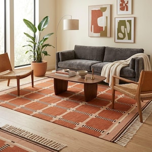 May include: A rectangular area rug featuring a terracotta orange and cream grid pattern with dark brown accents and fringe on the ends. The rug is positioned on a light wood floor beneath a dark gray sofa, a wooden coffee table, and two chairs.