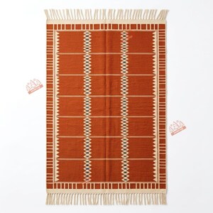 May include: A rectangular area rug in a deep burnt orange colour featuring a cream-coloured geometric border and two vertical central columns of diamond-shaped patterns. The rug is finished with a fringe of tassels along the top and bottom edges.