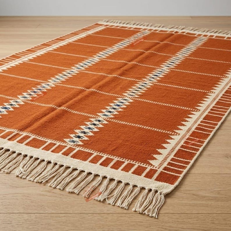 May include: A rectangular area rug in a deep burnt orange colour featuring a repeating pattern of white horizontal stripes and a central vertical band of dark blue and white geometric diamond shapes, finished with white fringe on the short ends.