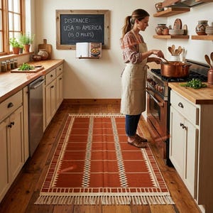 May include: A rectangular area rug featuring a burnt orange base with a white grid pattern and decorative fringe on the short ends. The rug is positioned on a wooden floor in a kitchen setting, showcasing a woven texture and geometric border details.