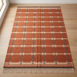 May include: A rectangular area rug featuring a burnt orange base color with a grid pattern of cream-colored horizontal lines and black-and-white striped vertical lines. The rug is finished with cream-colored fringe along the top and bottom edges.