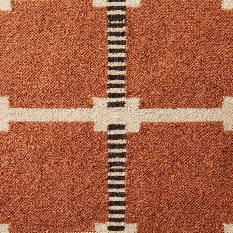 May include: A woven textile featuring a grid pattern of large burnt orange squares separated by thin white horizontal lines and vertical bands composed of alternating thin black and white stripes that intersect at the center of the grid.