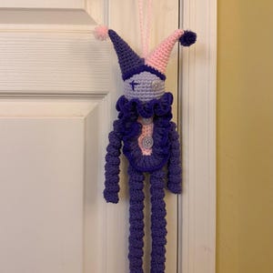 Op de afbeelding: Een handgemaakte gehaakte narrenpop met een tweekleurige paarse en lichtroze puntmuts met pompons, een lichtpaars gezicht met een kruissteek-oog en een knoop-oog, een donkerpaarse ruchekraag en lange paarse ledematen met knoopaccenten.