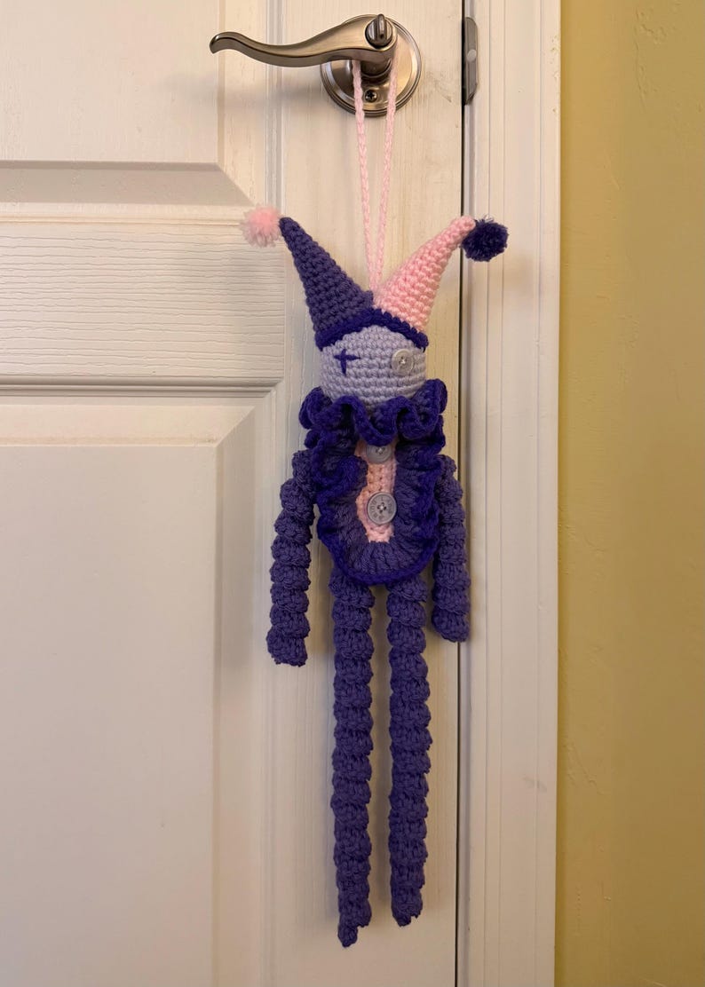 Op de afbeelding: Een handgemaakte gehaakte narrenpop met een tweekleurige paarse en lichtroze puntmuts met pompons, een lichtpaars gezicht met een kruissteek-oog en een knoop-oog, een donkerpaarse ruchekraag en lange paarse ledematen met knoopaccenten.