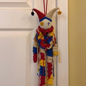 Op de afbeelding: Een gehaakte narrenpop met een cr&egrave;mekleurig hoofd, een blauw kruis en een geel bloemdetail als oog, met een rood, blauw en geel ruchekraagje. Het lichaam heeft lange, bungelende ledematen in afwisselende rode, blauwe en gele garensegmenten.