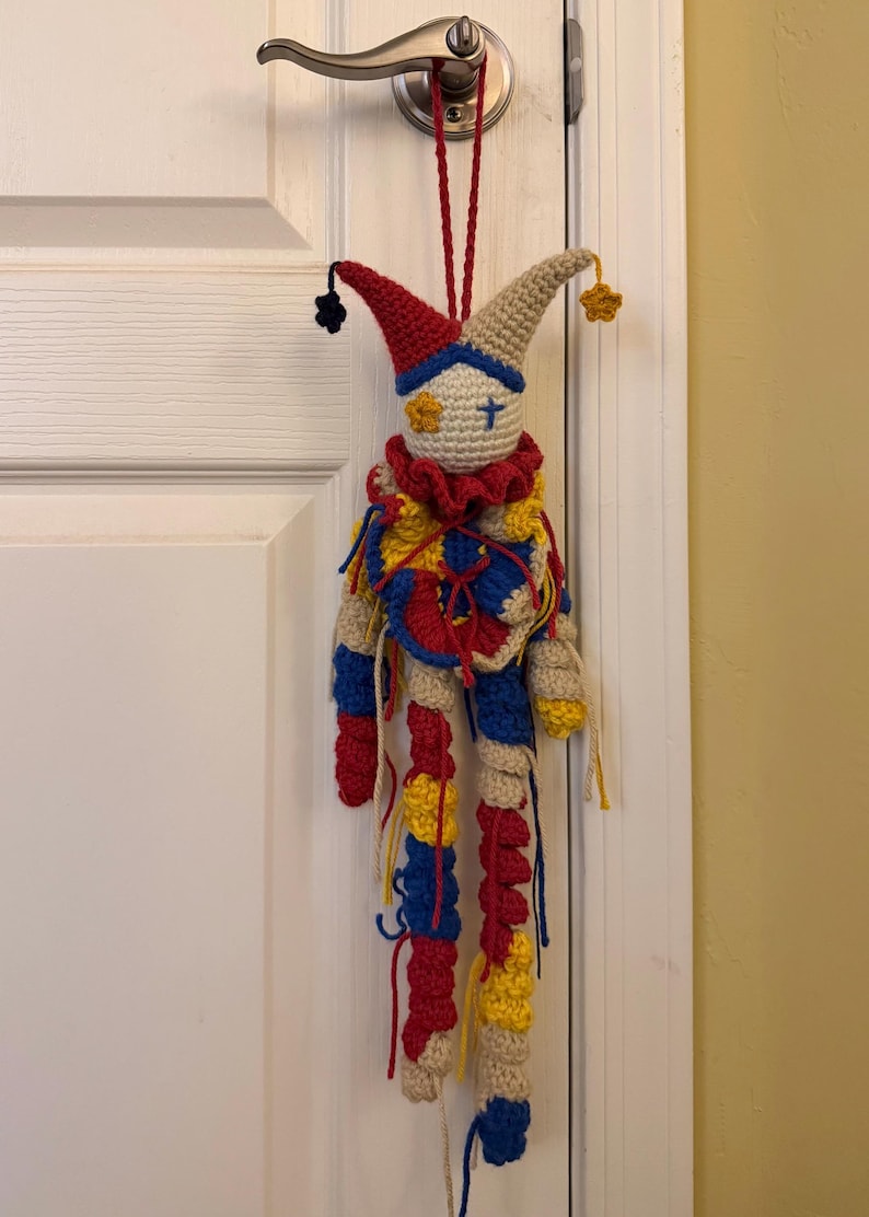 Op de afbeelding: Een gehaakte narrenpop met een cr&egrave;mekleurig hoofd, een blauw kruis en een geel bloemdetail als oog, met een rood, blauw en geel ruchekraagje. Het lichaam heeft lange, bungelende ledematen in afwisselende rode, blauwe en gele garensegmenten.