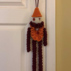 Op de afbeelding: Een handgemaakte gehaakte pop met een puntige oranje hoed, een cr&egrave;mekleurig hoofd met een rood knoop-oog en een gestikt kruis-oog, een geplooide oranje en donkerrode kraag, en lange, bungelende donkerrode armen en benen aan een lus.
