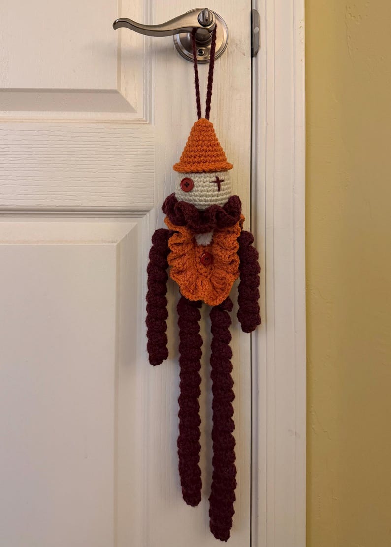 Op de afbeelding: Een handgemaakte gehaakte pop met een puntige oranje hoed, een cr&egrave;mekleurig hoofd met een rood knoop-oog en een gestikt kruis-oog, een geplooide oranje en donkerrode kraag, en lange, bungelende donkerrode armen en benen aan een lus.
