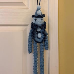 Op de afbeelding: Een handgemaakte gehaakte pop met een lichtblauw hoofd, kruissteekogen, een puntmuts met een pompon en een donkerblauwe rucheskraag. De pop heeft lange, textuurrijke blauwe armen en benen en een lus aan de bovenkant van de muts om op te hangen.