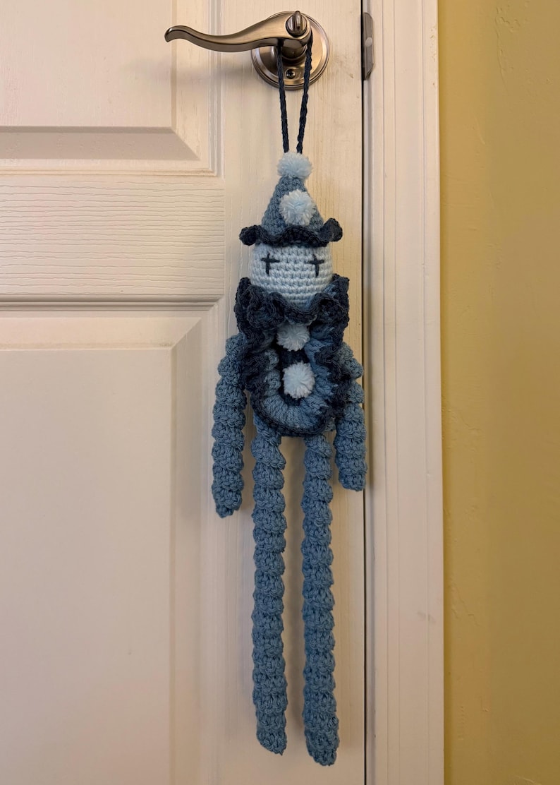 Op de afbeelding: Een handgemaakte gehaakte pop met een lichtblauw hoofd, kruissteekogen, een puntmuts met een pompon en een donkerblauwe rucheskraag. De pop heeft lange, textuurrijke blauwe armen en benen en een lus aan de bovenkant van de muts om op te hangen.