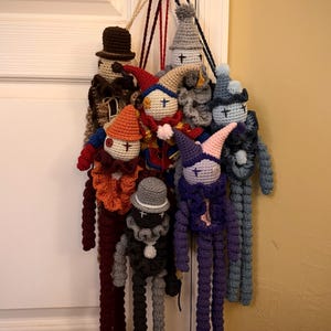 May include: A collection of handmade crochet dolls, each with unique features. The dolls have various hats, beards, and outfits in colors like brown, red, orange, gray, blue, and purple. They are hanging from a door handle.