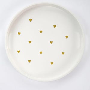 May include: A white, round serving tray with a raised edge. The tray is decorated with small, gold-colored heart shapes scattered across the surface. The tray is suitable for serving food or as a decorative piece.
