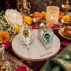 May include: Two silver-toned decorative pins, one shaped like a feather and the other like a leaf, featuring embedded crystals and a central colored stone. The pins rest on a round stone base with the text Shubh Diwali printed on it.