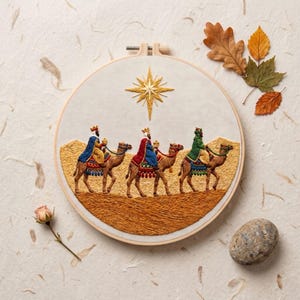 May include: A circular embroidery hoop featuring a needlepoint scene of three figures in crowns riding camels across a textured golden desert landscape under a large, stitched gold star. The figures wear blue, red, and green robes with detailed trim.