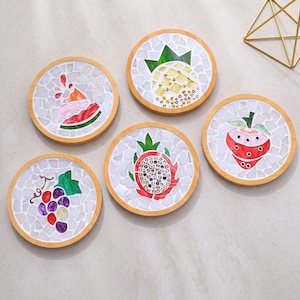 Op de afbeelding: Een set van vijf ronde onderzetters met lichte houten frames, elk voorzien van een uniek mozaïekontwerp van fruit gemaakt van witte en gekleurde glasstukjes. De ontwerpen omvatten een watermeloenpartje, een ananas, een drakenfruit, een aardbei en druiven.