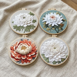 May include: Four circular wooden embroidery hoops featuring detailed floral needlework. Three hoops display white chrysanthemum flowers with green leaves on light fabric, while one hoop features a vibrant orange flower on a light background.
