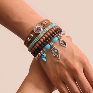 Könnte beinhalten: Ein mehrlagiges Armbandset mit braunen Wildlederbändern, türkisfarbenen Scheibenperlen und dunkelbraunen Holzperlen. Das Set enthält silberfarbene Metallanhänger in Form einer Blume, eines Blitzes und eines Blattes mit türkisfarbenen Akzenten.