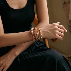 Puede incluir: Un conjunto de cinco pulseras en la muñeca con cuentas en tonos ámbar, dorado y crema. El set incluye cuentas redondas lisas, discos metálicos texturizados y piezas cilíndricas con patrones, creando un estilo de capas superpuestas.
