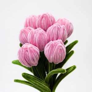 May include: A bouquet of artificial flowers crafted from pink pipe cleaners, featuring rounded, textured petals and long, dark green fuzzy leaves. The stems are bundled together, creating a dense arrangement of multiple individual flower heads.