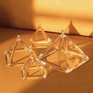 Puede incluir: Cuatro objetos decorativos transparentes en forma de pirámide de vidrio. Cada pirámide tiene una base cuadrada y una parte superior puntiaguda. Las pirámides varían en tamaño, siendo la más grande la del primer plano. Los objetos están sobre una superficie marrón.