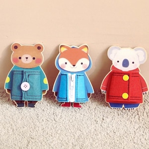 Peut inclure: Trois figurines en bois d'animaux : un ours, un renard et un koala. Chaque figurine porte un manteau en feutre. L'ours a un manteau bleu avec un bouton, le renard a un manteau bleu avec une fermeture éclair et le koala a un manteau rouge avec des boutons jaunes.