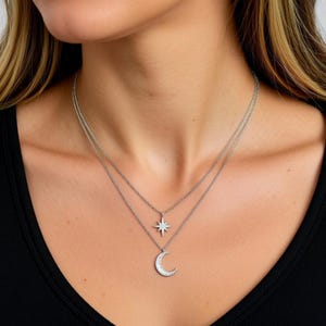 Può includere: Una collana d'argento a più strati con due ciondoli. Il ciondolo superiore è a forma di stella, e il ciondolo inferiore è a forma di mezzaluna. La collana è indossata su un top nero.