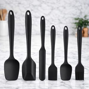 Könnte beinhalten: Ein Set aus sechs schwarzen Küchenutensilien aus Silikon, darunter ein Spatel, ein Schaber und ein Pinsel, auf einer Marmorarbeitsplatte. Jedes Werkzeug hat ein Aufhängeloch. Die Utensilien sind zum Kochen und Backen bestimmt.