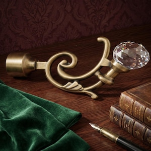 May include: A decorative curtain rod holder with a faceted crystal ball finial. The holder is made of brass-colored metal with an ornate, swirling design. A green velvet fabric and two leather-bound books are also in the image.