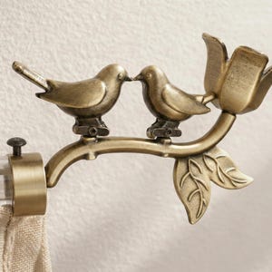 Puede incluir: Terminal de barra de cortina en tono bronce con dos pájaros posados en una rama con hojas y una flor. El terminal está unido a una barra transparente con una cortina beige. El diseño añade un toque decorativo.