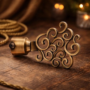Może przedstawiać: Brązowy zakończenie drążka do zasłon z ozdobnym, wirującym wzorem. Zakończenie ma cylindryczną podstawę ze śrubą i dekoracyjny, przypominający zwój wzór. Drążek jest umieszczony na drewnianej powierzchni.