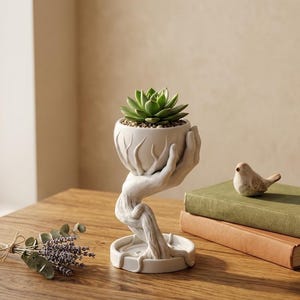 Może przedstawiać: Biała ceramiczna donica w kształcie dłoni trzymających sukulenta. Donica stoi na drewnianym stole obok małej figurki ptaka i dwóch książek. Na stole znajduje się również pęczek suszonej lawendy.