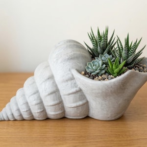 Könnte beinhalten: Ein Set aus drei Keramikbechern mit gesprenkelter, matter Oberfläche. Jeder Becher hat eine andere Farbe: Hellblau, Creme und Grau. Die Becher haben ein schlichtes, modernes Design und einen bequemen Griff.