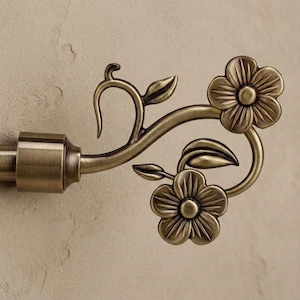 Peut inclure: Extrémité de tringle à rideau de couleur bronze avec un motif floral. La décoration comprend deux fleurs et des feuilles détaillées, avec une tige incurvée. La tige elle-même est de forme cylindrique et de finition bronze assortie. Le design est orné et décoratif.