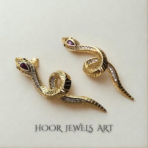 Può includere: Due gioielli dorati a forma di serpente. Ogni serpente ha un occhio color rubino ed è adornato con piccole pietre preziose trasparenti. I gioielli sono esposti su una superficie di colore chiaro. Il testo "HOOR JEWELS ART" è visibile.