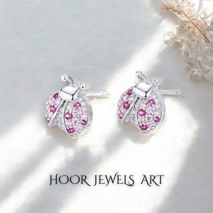 Pode incluir: Um par de brincos de botão prateados em forma de joaninha. O corpo da joaninha é cravejado com pedras preciosas rosa e transparentes. Os brincos têm fecho de pino e pressão. O texto "HOOR JEWELS ART" está na parte inferior da imagem.