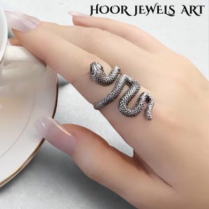 Puede incluir: Un anillo plateado con forma de serpiente que se enrolla alrededor de un dedo. El anillo presenta un diseño detallado de serpiente con escamas texturizadas y un pequeño ojo oscuro. El texto "HOOR JEWELS ART" está en la parte superior.