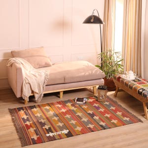 Puede incluir: Una alfombra tejida rectangular con un patrón repetitivo de formas de cruz blancas sobre rayas verticales en tonos naranja, rojo, azul y verde. La alfombra tiene bordes con flecos y el texto wovenrugz está impreso en blanco sobre la superficie.