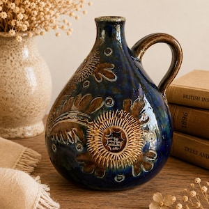 Pode incluir: Uma jarra de cerâmica azul-escura com corpo arredondado e uma única alça. A superfície apresenta padrões florais e motivos circulares em relevo na cor castanha. O esmalte tem um acabamento brilhante com variações de cor no exterior.