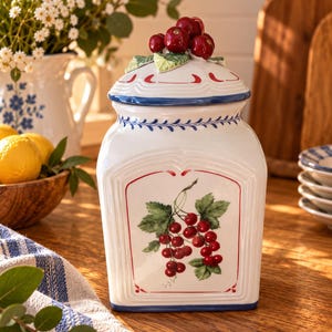 Könnte beinhalten: Ein weißer Keramikbehälter mit quadratischem Boden und abgerundeten Ecken, der auf der Vorderseite ein handgemaltes Motiv von roten Johannisbeeren zeigt. Der Deckel ist mit einer skulptierten Gruppe roter Kirschen und grüner Blätter verziert.