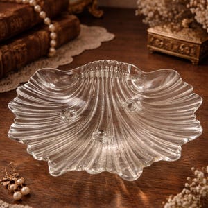 Könnte beinhalten: Eine durchsichtige, muschelförmige Glasschale mit gewelltem Rand. Die Schale hat drei kleine, kreisförmige Füße und liegt auf einer Holzoberfläche. Die Schale ist von anderen dekorativen Gegenständen umgeben.