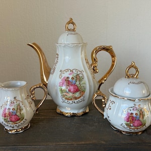 May include: A white porcelain tea set with gold accents. The set includes a teapot, creamer, and sugar bowl, each adorned with a colorful pastoral scene. The teapot and sugar bowl have lids. The set is displayed on a dark wooden surface.