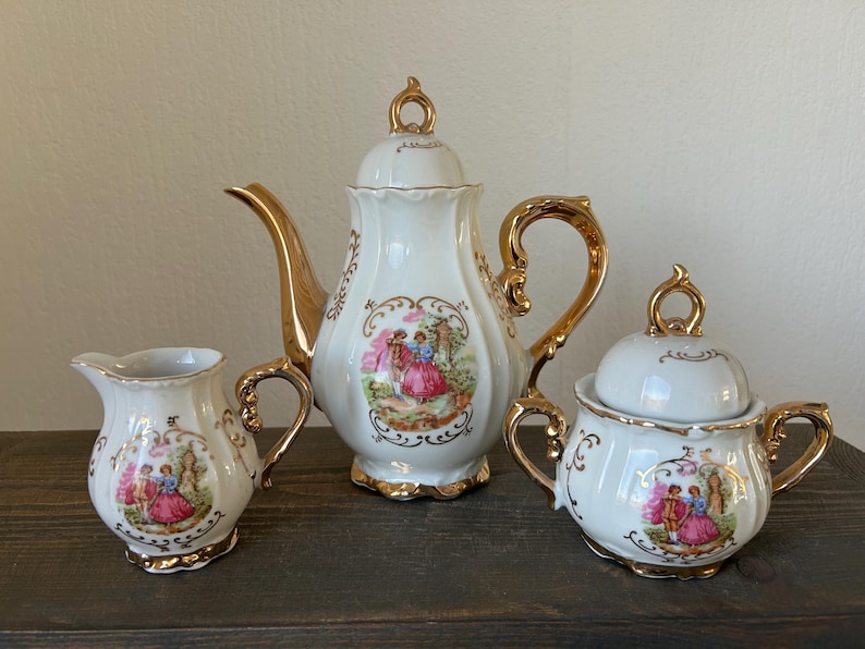 May include: A white porcelain tea set with gold accents. The set includes a teapot, creamer, and sugar bowl, each adorned with a colorful pastoral scene. The teapot and sugar bowl have lids. The set is displayed on a dark wooden surface.