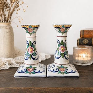 May include: A pair of white ceramic candle holders featuring a hand-painted floral motif in pink, green, and blue. The square bases and cylindrical stems are decorated with intricate scrollwork and a yellow lattice pattern at the top rim.