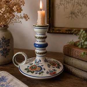 Może przedstawiać: Kremowy ceramiczny świecznik z uchwytem, ozdobiony ręcznie malowanymi niebieskimi i zielonymi poziomymi paskami na trzonku oraz kolorowym kwiatowym wzorem w odcieniach pomarańczu, błękitu i zieleni na podstawie, z zapaloną białą świecą.