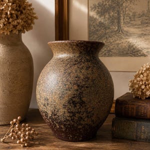 Könnte beinhalten: Eine runde Keramikvase mit weiter Öffnung und einer texturierten, gesprenkelten Oberfläche in Dunkelbraun, Beige und Schwarz. Das Gefäß weist ein fleckiges Muster auf, wobei der dunklere Boden in einen helleren, erdigen Ton zum Rand hin übergeht.